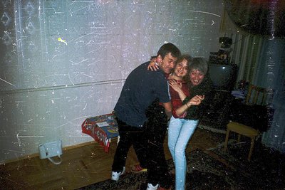 Vintage indoor party scene with three adults hugging, surrounded by confetti. The man wears a dark sweater, woman in jeans an...