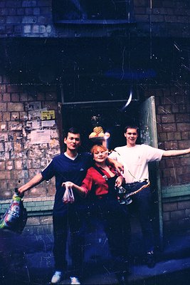 Three young men pose outside a brick building in a 1970s-style photo. Left: dark sweater, holding a green bottle. Center: red...