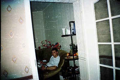 Vintage interior shot of a man seated on a patterned sofa in a dimly lit room, holding a book. Reflections in a glass door re...