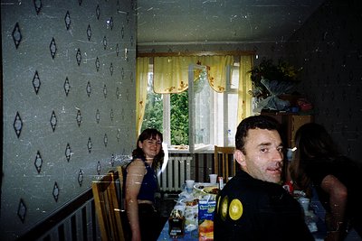 Vintage indoor dining scene with geometric wallpaper and floral curtains. Two adults seated at a table with bottled drinks, a...