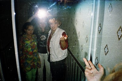 Vintage elevator interior with geometric tile patterns, featuring three individuals—one pointing toward the camera. The centr...