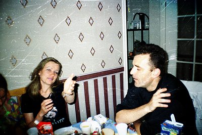 Two individuals seated indoors at a table with retro patterned wallpaper (diamond motifs). The man gestures with his hand whi...