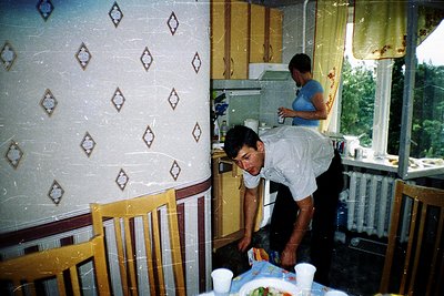 Vintage kitchen scene featuring two men in casual attire (one in a white apron) preparing food. Patterned wallpaper with diam...
