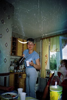 Mid-20th-century kitchen interior with textured ceiling and floral-patterned wallpaper. Woman in light blue apron pours from ...