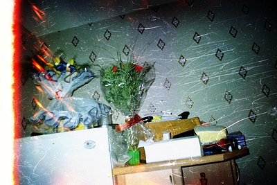 Vintage-style photo of a cluttered indoor corner with peeling wallpaper featuring geometric patterns. A floral arrangement in...