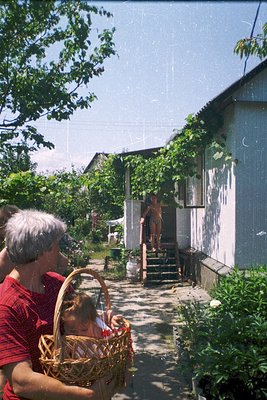 Vintage snapshot of a rural courtyard with a woman in red holding a basket of produce, likely tomatoes, while a child stands ...