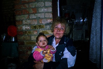 Vintage indoor scene featuring an elderly woman holding a young child in a pink/yellow outfit. Brick wall and vintage applian...