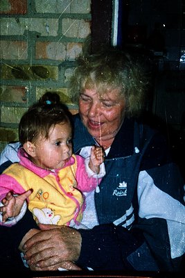 A tender intergenerational moment captured: an elderly woman in a vintage **Raidr** jacket cradles a baby in a pastel onesie,...