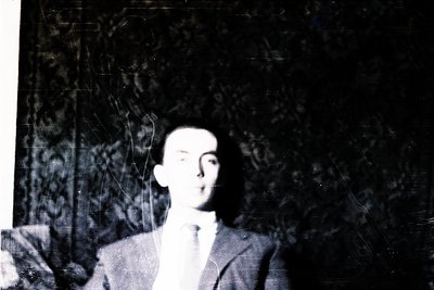 Blurred black-and-white portrait of a man in formal attire—dark suit, white shirt, and tie—posing indoors against a patterned...