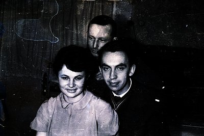 Vintage black-and-white portrait of three individuals indoors, likely mid-20th century. The woman in the foreground wears a s...