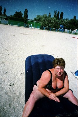 Vintage beach scene with woman sitting on a blue inflatable mat, mid-20th century attire. Green fence and trees in background...