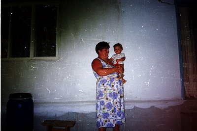 A woman in a floral dress cradles an infant indoors, likely in a rural or modest home setting. The faded wall and wooden furn...