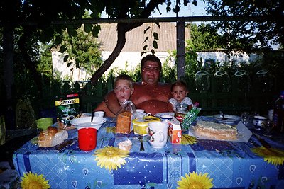 Family meal under a shaded outdoor canopy, likely late 20th century. Three generations seated at a table adorned with sunflow...