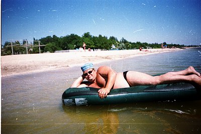 Vintage beach scene featuring a man reclining on a green inner tube in shallow water, wearing sunglasses and a swim cap. Sand...