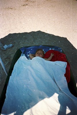 Aerial shot of an infant sleeping in a makeshift bed under a large rock, wrapped in a blue blanket with red accents. The scen...