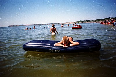 Vintage seaside scene featuring a person resting on an "INTEX" inflatable raft in shallow water. Crowded beach with swimmers,...