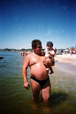 A man in swim trunks holds a baby in a striped hat at a beach, mid-20th century. Water splashes around them, and the shore is...