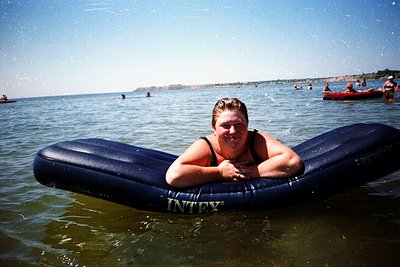 Woman reclining on a blue **Intex** inflatable raft in shallow coastal waters, smiling at the camera. Background shows distan...
