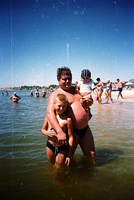 A man carries a child in shallow seaside water, with another child clinging to his waist. Mid-20th century beach attire—swim ...