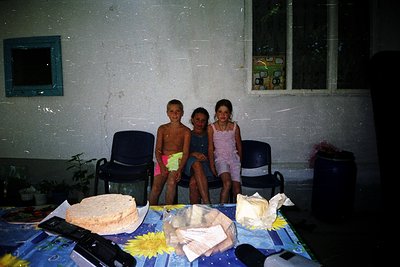 Three children pose indoors on blue plastic chairs, seated at a table with paper plates and a cake. The boy wears a pink clot...