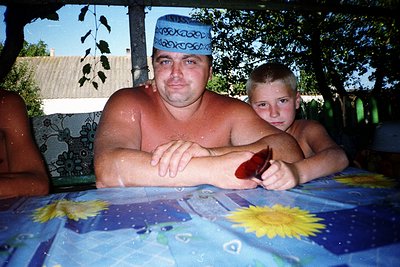 A man and boy sit at an outdoor table under a shaded canopy. The man wears a traditional embroidered cap and a patterned, wet...