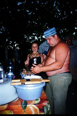 A man and child preparing food outdoors under trees, likely at a rural gathering. The man wears a traditional headscarf and b...