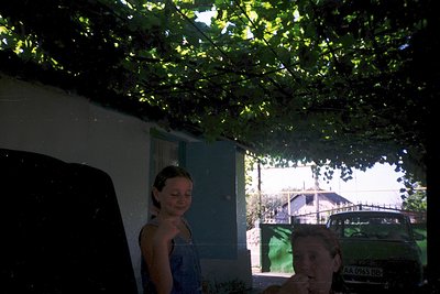 Vintage-style photo of two people under dense foliage, likely a grapevine pergola. Woman in sleeveless top, man in dark shirt...