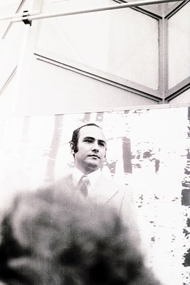 Black-and-white portrait of a man in a suit and tie, framed by a large, reflective window showing urban architecture. Mid-cen...