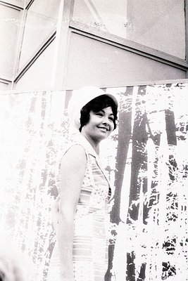 Black-and-white portrait of a woman in a floral-patterned dress, posed indoors with a curtain backdrop. Her short hair and vi...