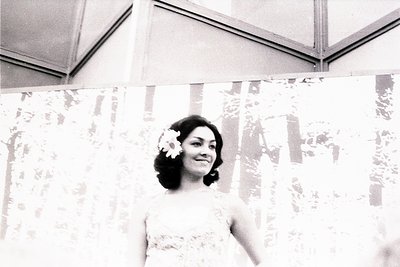 Black-and-white portrait of a woman in a formal gown with floral headpiece, set against a draped backdrop of floral-patterned...