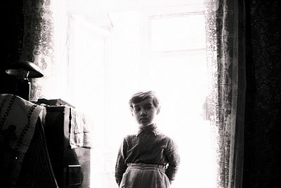 Young boy in early 20th-century attire stands in dimly lit interior, sunlight streaming through lace curtains. Wooden furnitu...