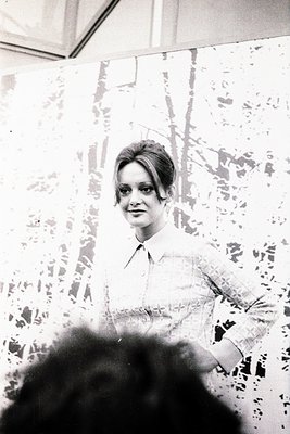Vintage black-and-white portrait of a woman in a 1960s-style blouse, standing indoors against a textured wall. Her short, vol...