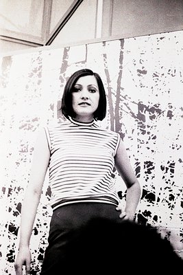 Portrait of a woman in a sleeveless striped top and high-waisted trousers, standing indoors against a patterned wall. Her sho...