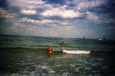 Vintage seaside scene with three swimmers in choppy waves under dramatic cloud cover. Film grain and faded tones suggest mid-...