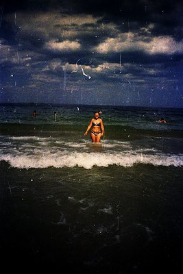 Vintage black-and-white seaside scene with lone swimmer in a two-piece bathing suit standing in shallow waves under a stormy ...
