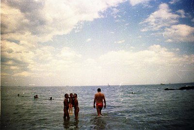 Vintage seaside scene with four people wading in shallow water, likely mid-20th century. Bright sunlight reflects off the oce...