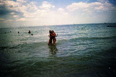 Vintage seaside embrace in shallow waves, mid-20th century beach vibes. Couple hugging near shore with distant swimmers. Soft...