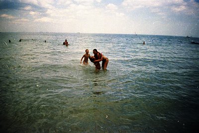 Vintage seaside scene: adult and child play in shallow waves, surrounded by other swimmers. Soft focus suggests mid-20th cent...
