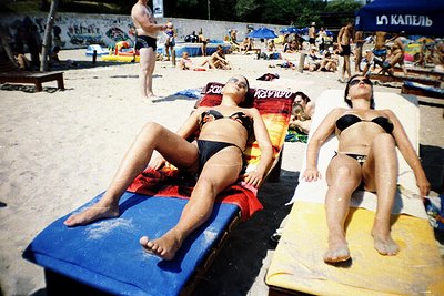 Sunbathing scene at a beachside resort, likely Eastern Europe, mid-20th century. Two women in black bikinis relax on lounge c...