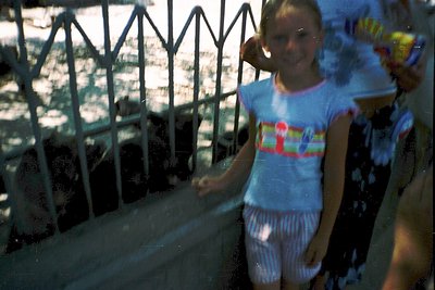 Vintage-style photo of a young girl in a light blue tie-dye shirt and striped shorts, standing on a wet concrete ledge beside...