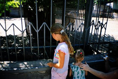 Two young girls in summer attire stand near a wrought-iron fence, one holding a rainbow-striped drink container. The older gi...