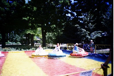 Vintage bumper boat ride in a park, featuring three white/blue pedal boats on a colored water track. Sign in Cyrillic ("ЛОДОЧ...