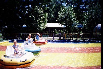 Vintage water bumper boats in a shallow, multi-colored pool with yellow, red, and blue lanes. Three children in 1970s-style s...
