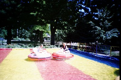 Vintage bumper boats in a shallow, multi-colored pool under evening tree canopy. Two riders in bright swimwear navigate red a...