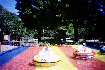Vintage bumper boat ride in a shaded outdoor pool area, featuring two inflatable boats (yellow & red) with "NIF" branding. A ...