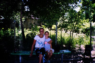 A woman and child pose together on a green metal bench in a lush, shaded forest setting. The woman’s white sleeveless top fea...