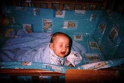 Vintage-style photo of an infant peacefully sleeping in a crib adorned with blue-and-white cartoon characters. Soft lighting ...