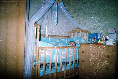 Vintage-style wooden crib with blue canopy and matching bedding, featuring a sleeping infant. Surrounding shelves hold plasti...