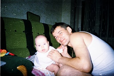 Intimate indoor portrait of a man and infant in a mid-20th-century setting. The man, shirtless in a white tank top, kneels be...