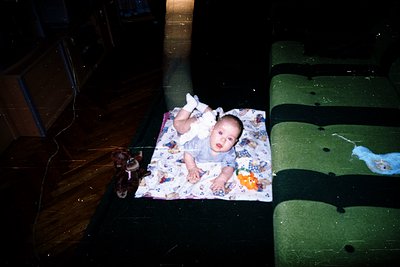 Vintage candid of infant lying on patterned blanket in dimly lit interior, surrounded by worn furniture. Green armchair and d...
